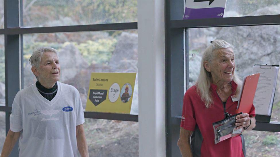 The twins listening to someone speaking off camera to the right. Jennifer is laughing while holding a clipboard. She is wearing a red sports top, while her sister has a white top on.