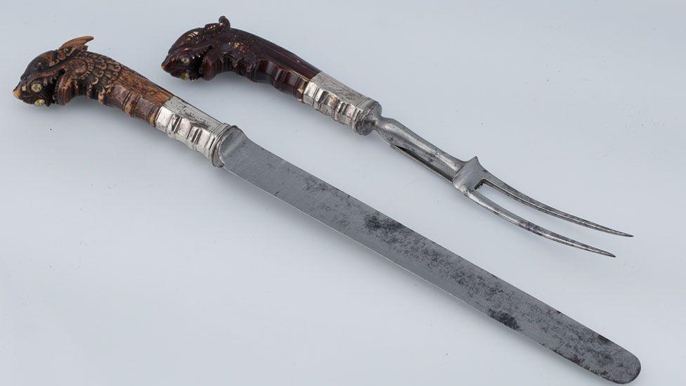 A 17th Century carving knife and two-pronged fork. The knife's handle is carved with a dragon-shaped head with pointed ears.