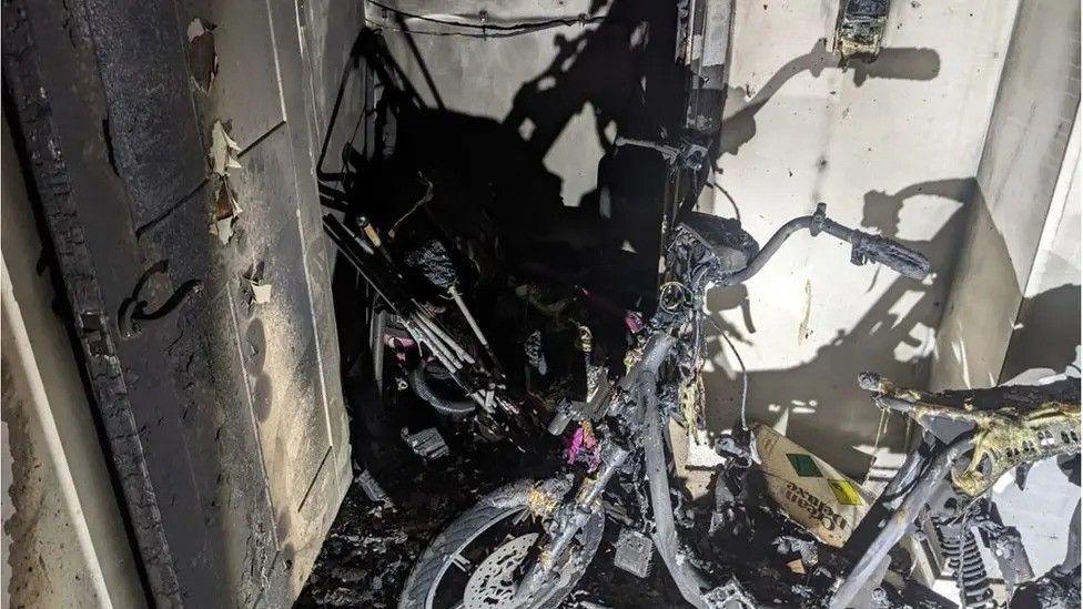 The charred aftermath of an e-bike fire in a confined space. The frame of the bike is severely burned, with melted components and blackened walls around it. Debris and scorched materials are scattered on the floor.