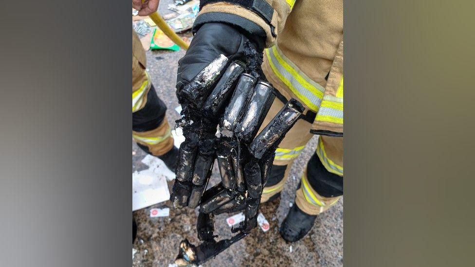 A set of lithium batteries that are charred and blackened by fire. They are being held by a firefighter who is wearing beige protective clothing with yellow and silver reflective strips