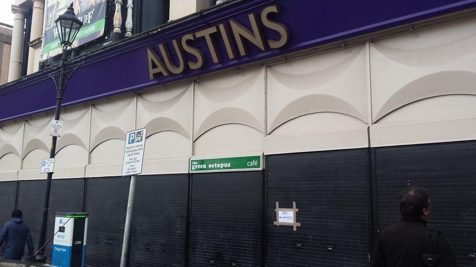 shutters pulled down on a store front. Above the closed shutters is a sign saying Austins. A lone shopper walks by