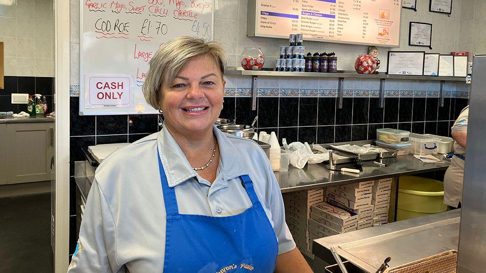 Sharon Foy owner of Sharon's Plaice chip shop stood behind the counter looking at the camera wearing a blue pinafore 