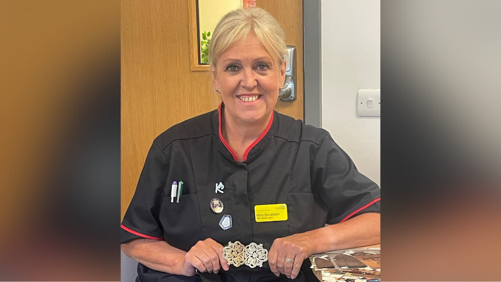 Nurse Mary Donaldson pictured in the hospital wearing a black uniform with red lined around the sleeves and collar. She has a purple and green pen in the breast pocket of the scrubs, yellow name tag, blonde hair tied back and is smiling at the camera. She is also holding a heirloom gold and silver buckle in her hands.