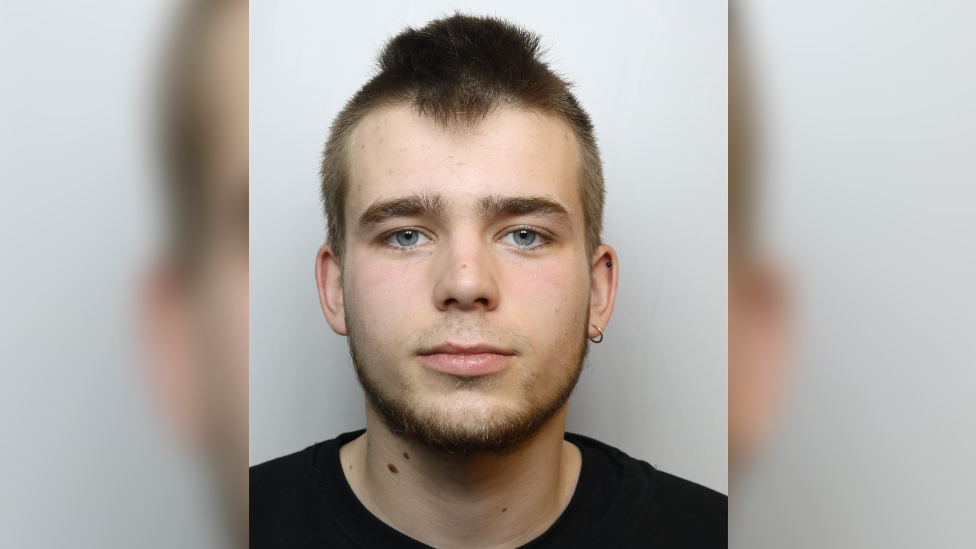 Louis Tavener head and shoulders photo taken after his arrest. He is wearing a black t-shirt, has brown hair, blue eyes and an earing in his left ear.