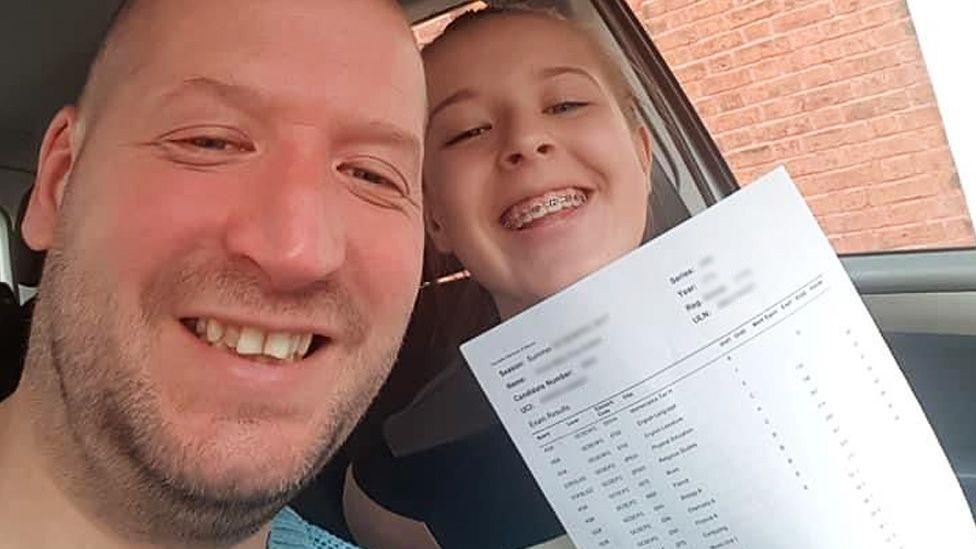 Anna smiles as she holds up her A-level results, printed on a white sheet of paper. She is sitting next to her dad, Les Sutton, in a car