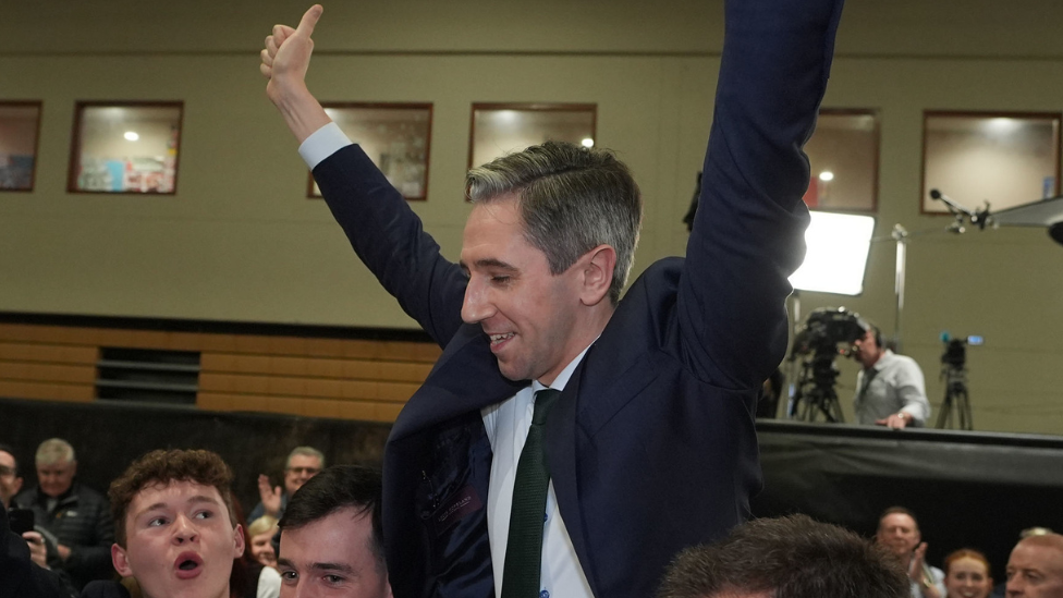 Simon Harris in blue suit jacket, raising arms in celebration as he is reelected, crowd holding him in the air