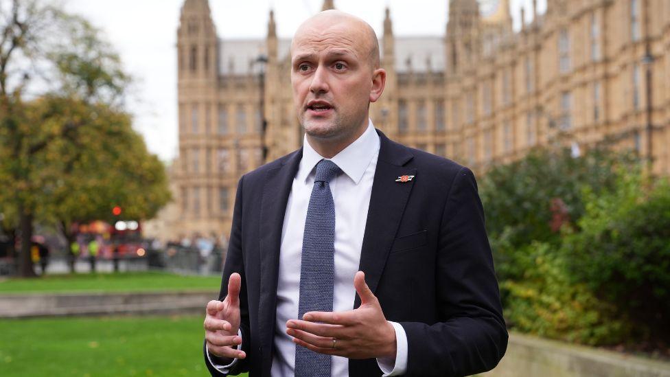 A bald man with a dark suit and blue tie speaks in front of the House of Parliament  