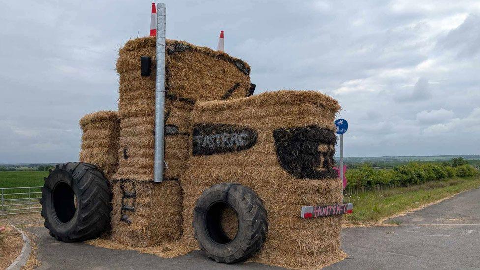 A straw bale sculpture showing a tractor. It has very fat black tyres resting on its side and two orange and white cones resting on its top. Its cab has a window highlighted in black and beneath it is a licence plate saying HuntsYF in pink. It is in a farm gate.