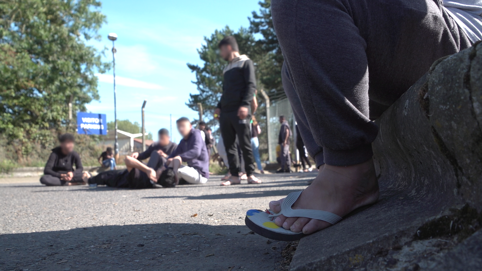 The photo is taken from road level. In the foreground is a person's legs, showing their feet in flip flops. They are sitting down. In the background a group of people are huddled, some sitting on the Tarmac.