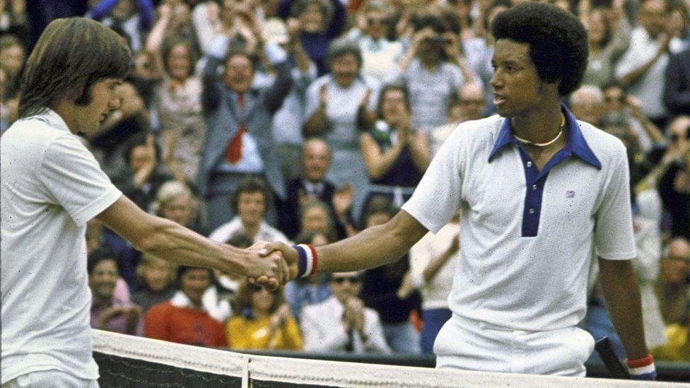 Jimmy Connors and Arther Ashe - both in tennis whites - shake hands over the net after Ashe's victory in the Wimbledon final of 5 July 2025. The clapping crowd can be seen behind them