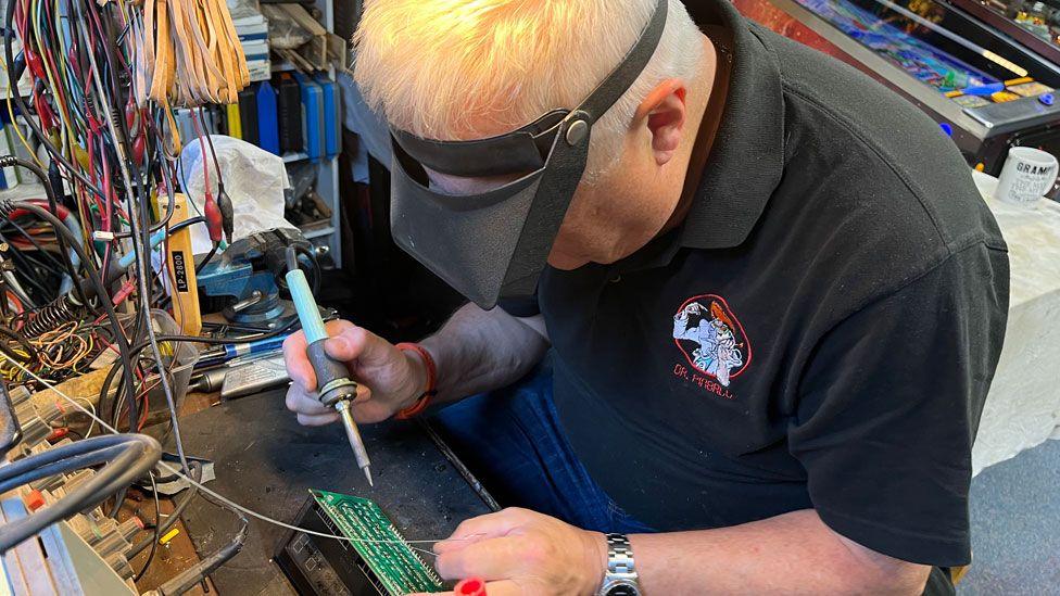 Mark Squires with short white hair, wearing a grey polo shirt, wearing an eye shield and holding a soldering iron in his right hand while looking down at an electronic board on a workbench