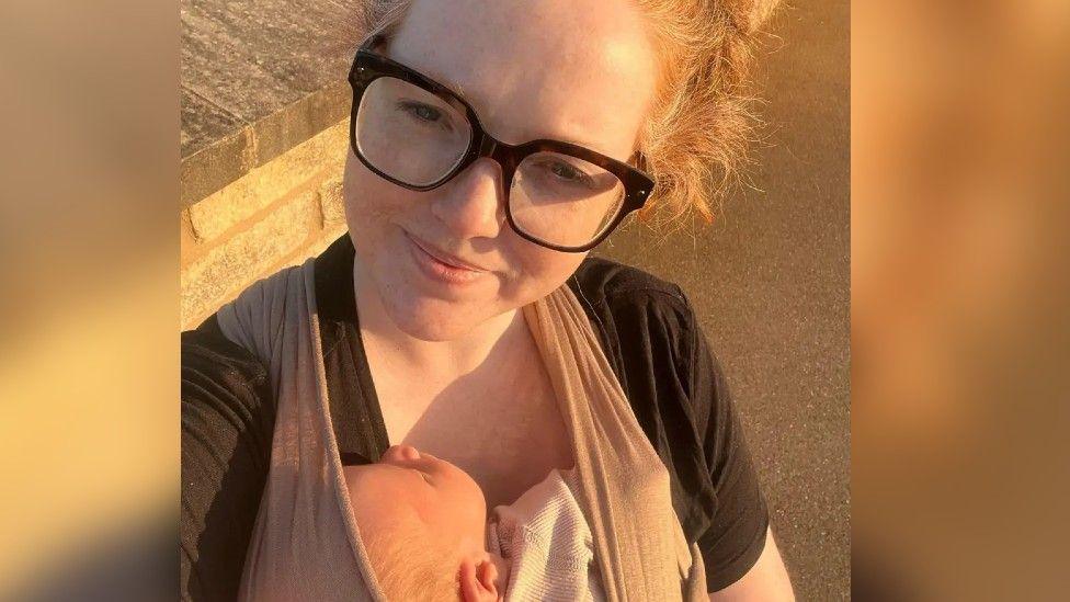 A woman with blonde hair and black glasses smiles at the camera. She is carrying a baby in a sling on her front.