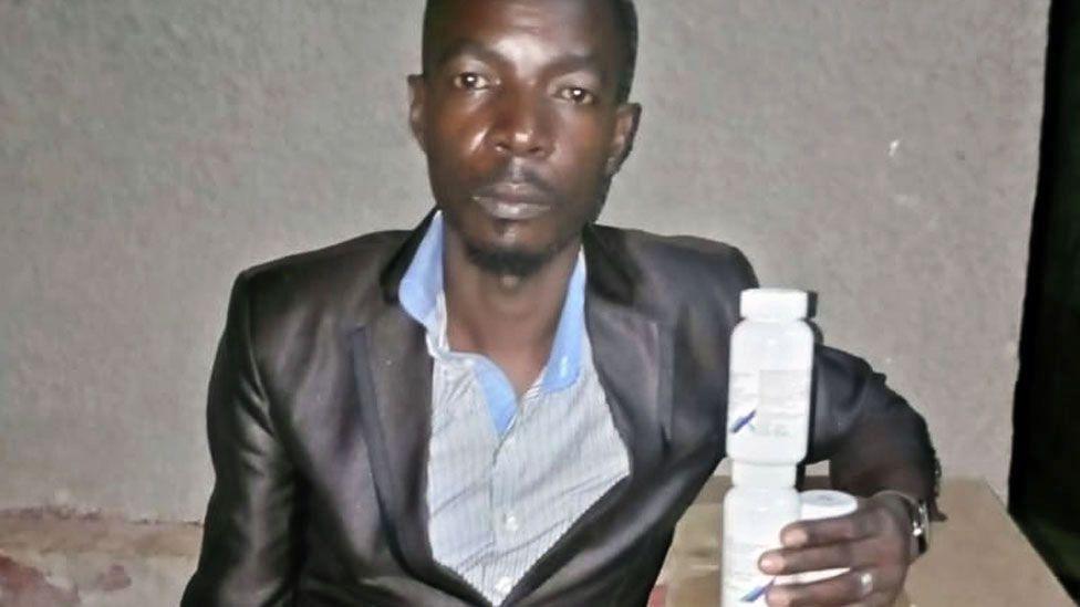 Mike Elvis Tusubira looking directly at the camera, wearing a black suit jacket and striped blue shirt. In his left hand he is holding some white plastic medication containers.