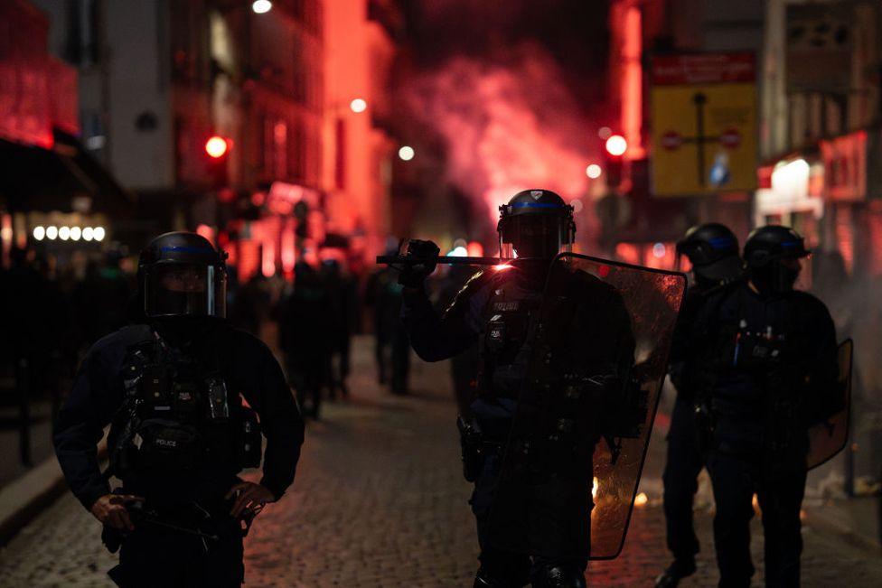 Police used tear gas to clear demonstrators at Place de la Republique in Paris, France during a rally on 30 June 2024.  Police in riot uniform stand in the foreground as red light an smoke rises up behind them at night.