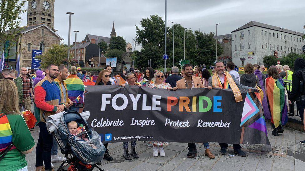 A group of people hold a foyle pride banner as they stand outside Derry's railway station, with a large crowd behind them. A number of buildings can be seen in the background while a man and a baby in a pram are standing to their right