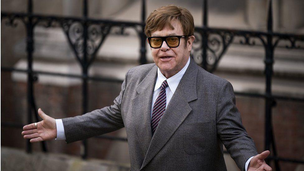 Elton John with short dark hair and black-framed glasses wearing a grey jacket, white shirt and striped tie, with his arms held out. He is standing in front of some black railings.