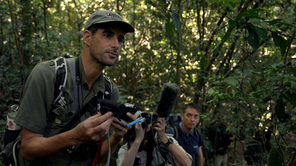 man in khaki shirt holds up recording equipment in the forest