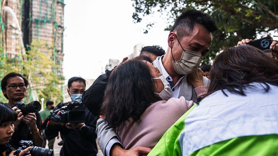 A man and a woman wearing face masks cling to each other, with photographers and the damaged tower blocks in the background