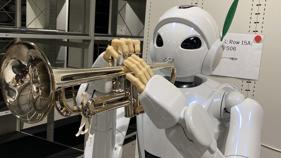 A white robot with large eyes and slightly human-like, with fingers holding a trumpet.