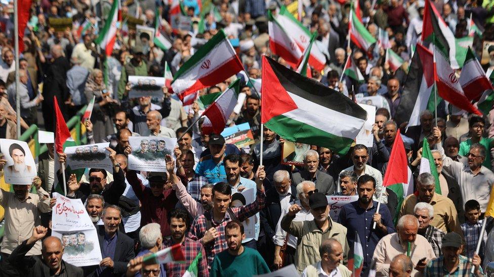 A large group of people protest in Iran, holding signs and Iranian flags