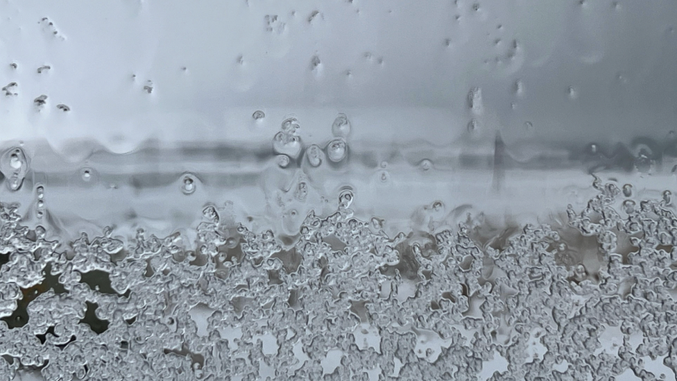 Photo of a window pane that has sleet accumulating. In the blurred distance it looks like white snow covers the ground