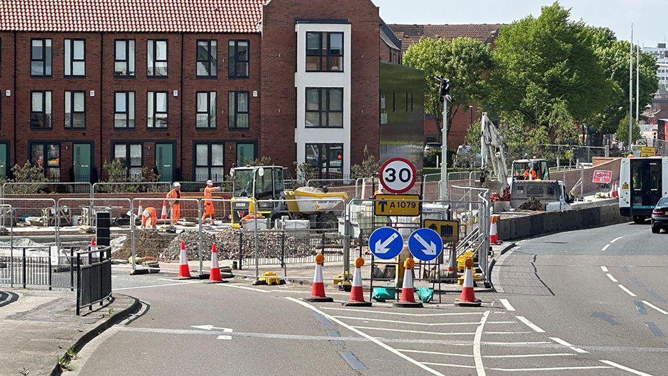 A junction with barriers traffic cones and signs. There are workers in the background wearing orange, high-visibility clothes, standing by a piece of machinery.