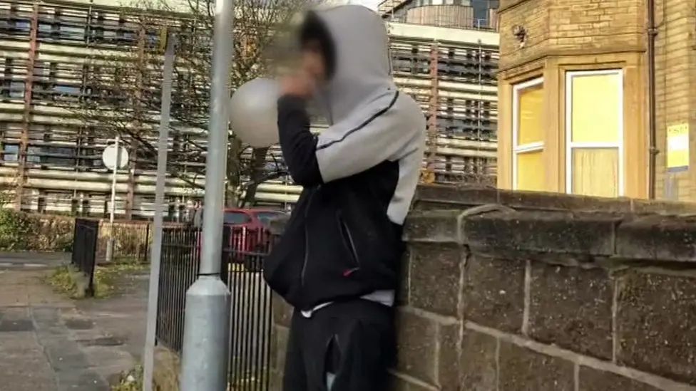 A young person wearing a hooded top and tracksuit bottoms, standing against a wall on a street corner and inhaling from a balloon. Their face is pixelated.