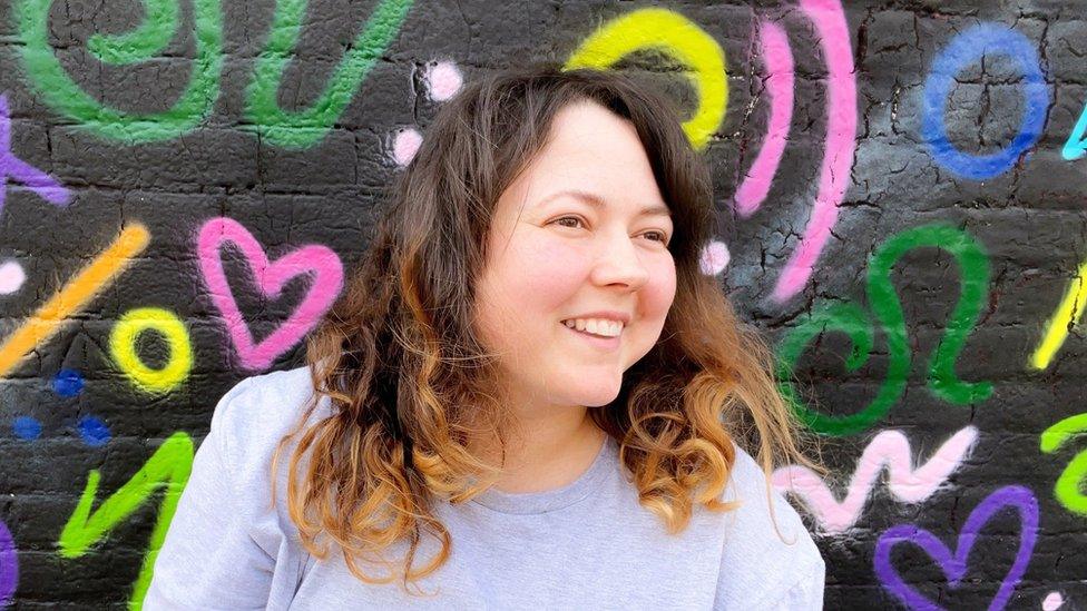 Amber, with dark curly hair, smiles as she leans against a black wall covered in multi-coloured symbols drawn in graffiti style