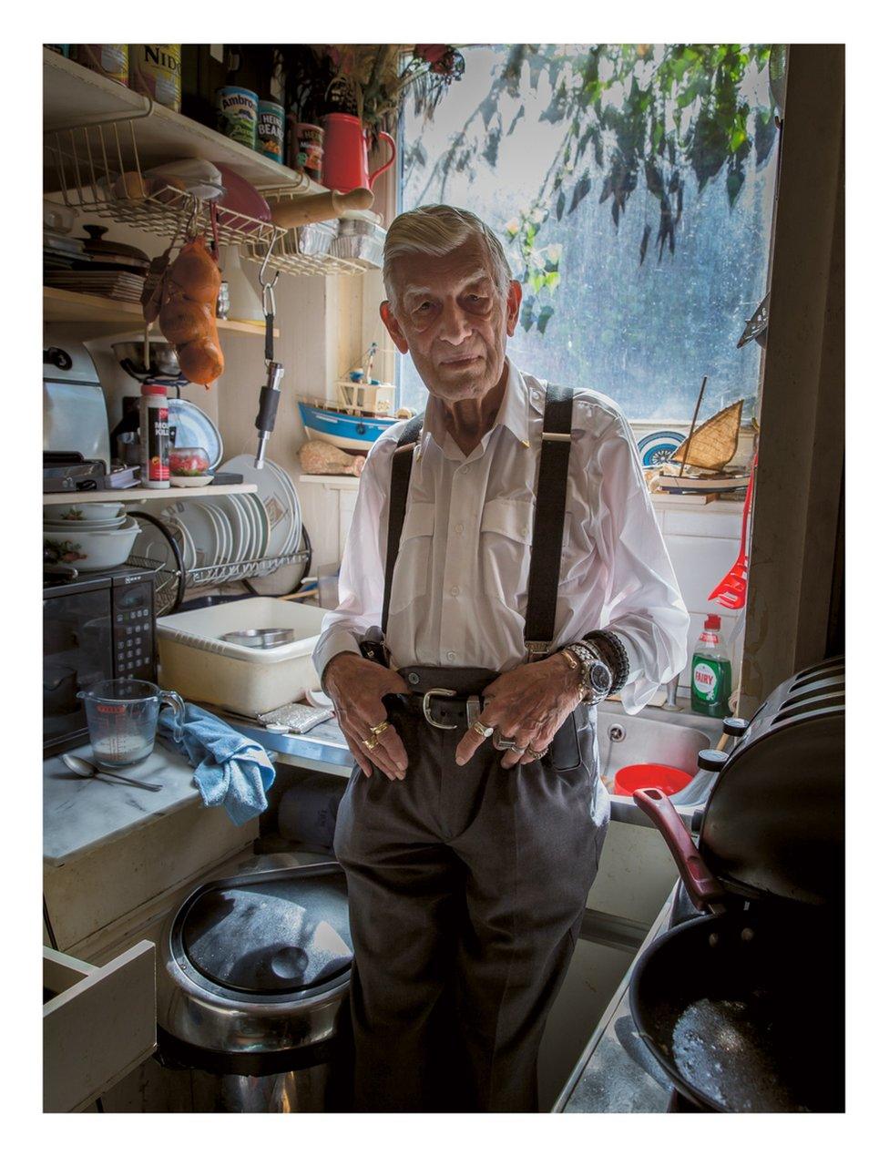 An elderly man stands in his kitchen