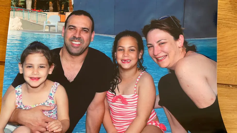 A family of four sit in front of a swimming pool. The man holds a young girl on his lap, an older girl sit next to him, with his wife on the right of the picture leaning in.