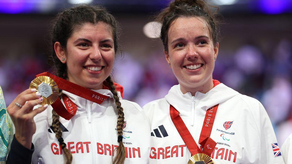 Khan Dannielle and Elizabeth Jordan holding their gold medals at the Paralympics in Paris. The pair are looking just away from the camera, and are wearing white ParalympicsGB tops. 