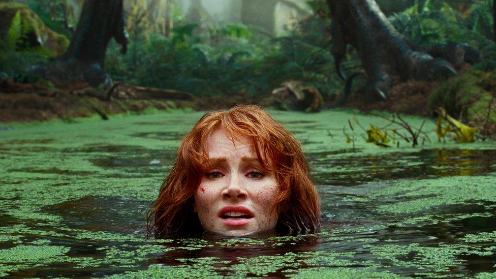 Bryce Dallas Howards is submerged in a swamp with a large dinosaur behind her.