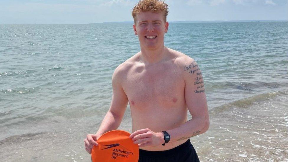 Louis Alexander standing on the shore holding an Alzheimer's Research UK orange swim cap