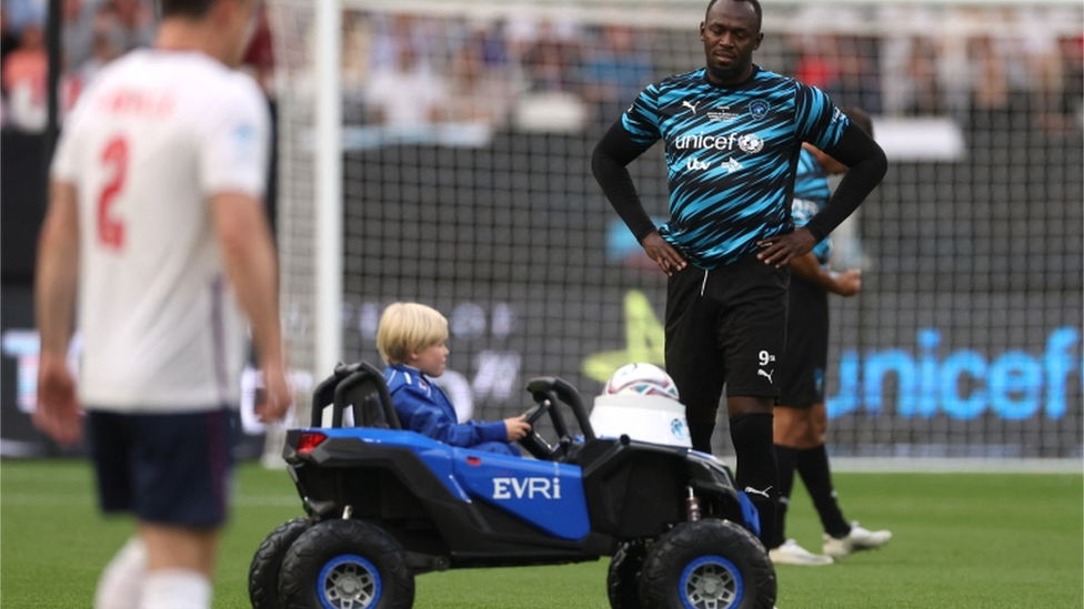 A young boy in a toy car delivers the ball onto the pitch as World XI"s Usain Bolt looks on before the match