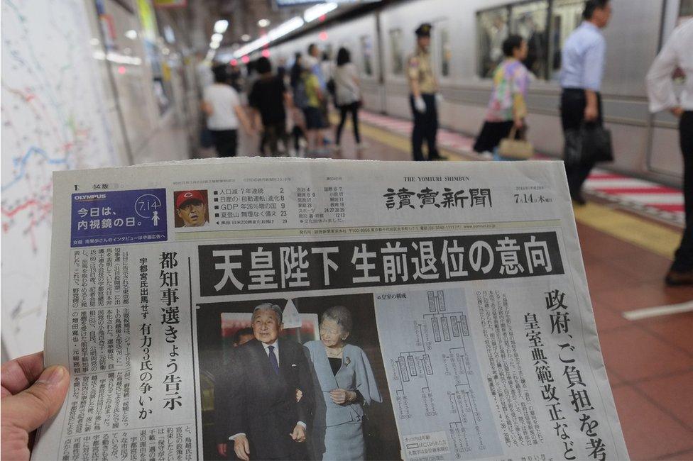 A Japanese newspaper with a story about reports the Emperor might abdicate on its front page, being held up for the camera in front of a view of people boarding a metro train in Tokyo. 14 July 2016.