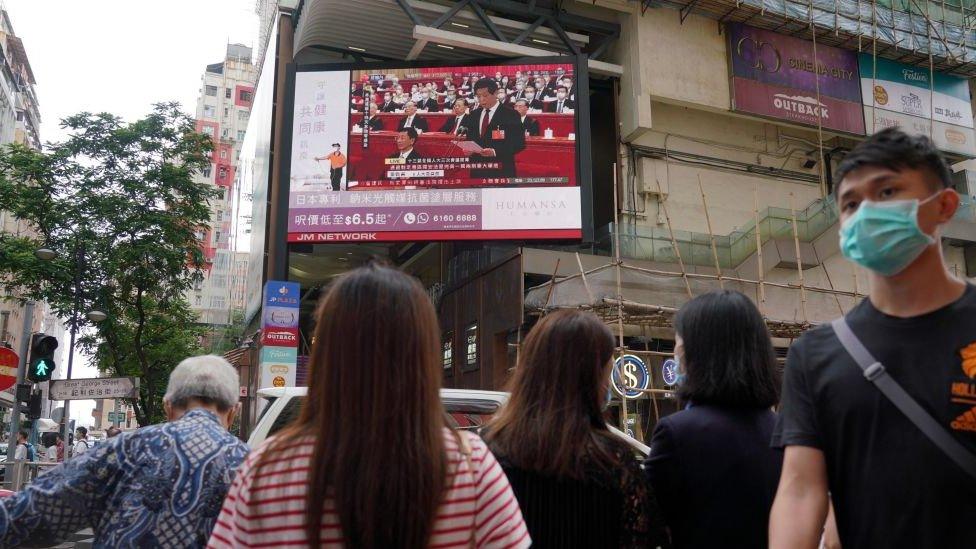 People wearing face masks walk past a large screen in Hong Kong displaying a live broadcast in the street of the National Congress