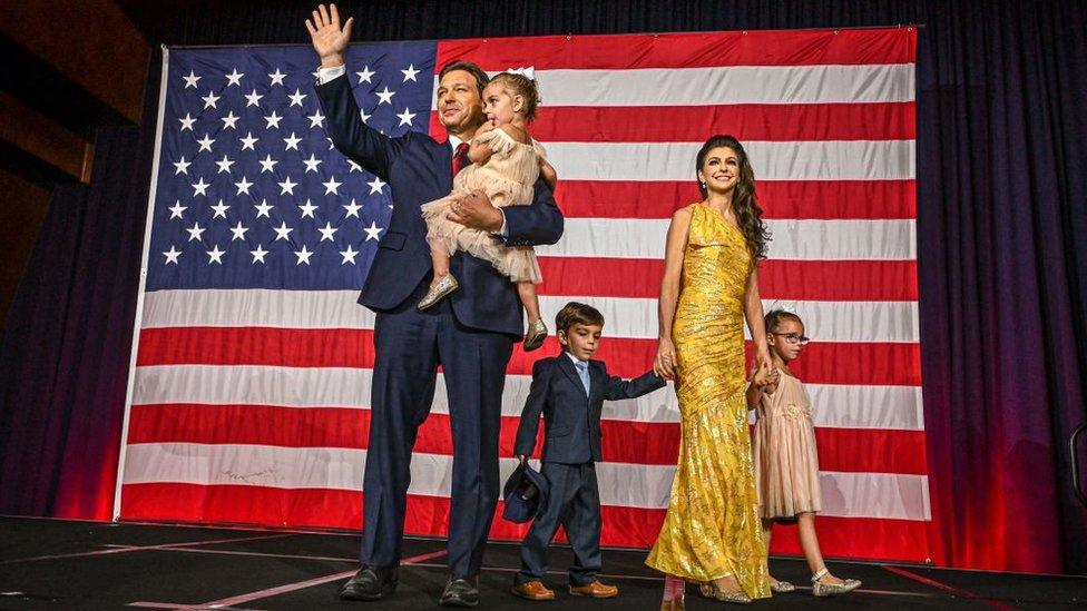 Ron DeSantis, his wife, Casey, and their three children pose in front of a United States flag. Mr Santis, wearing a blue suit with a white shirt and red tie, holds one of his young daughters, waving with his free arm. Mrs Desantis, wearing a fitted gold/yellow floor-length dress, stands with the couple's young son and daughter at her side, holding their hands.