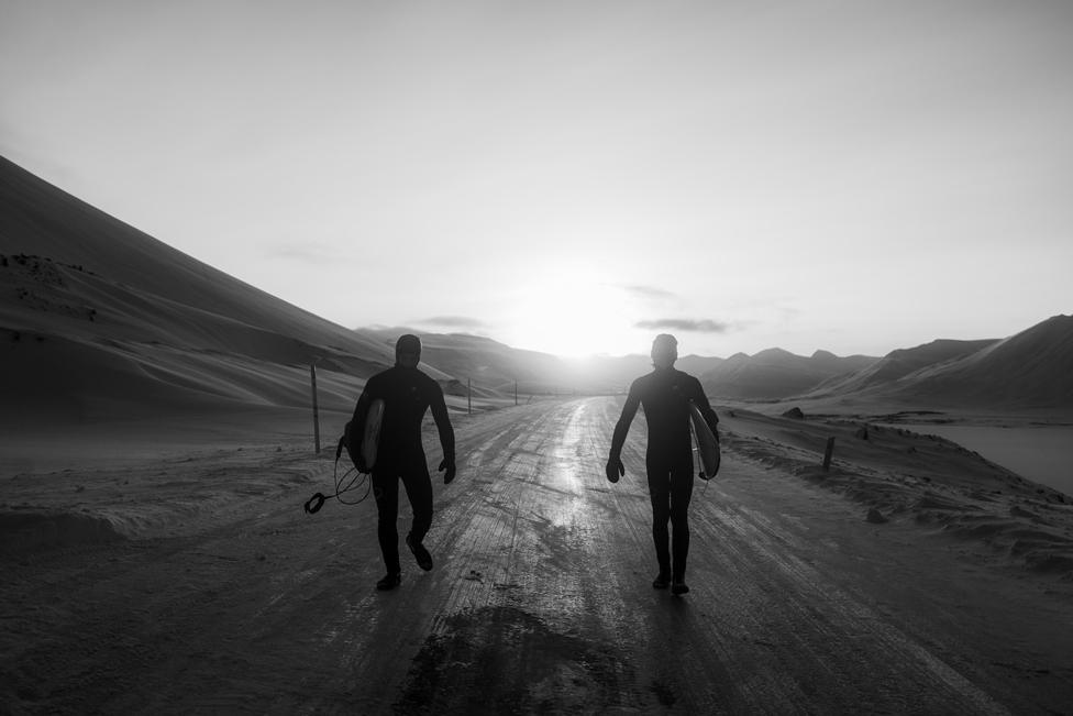 Surfers wearing wetsuits and holding surfboards walk along a path in Iceland