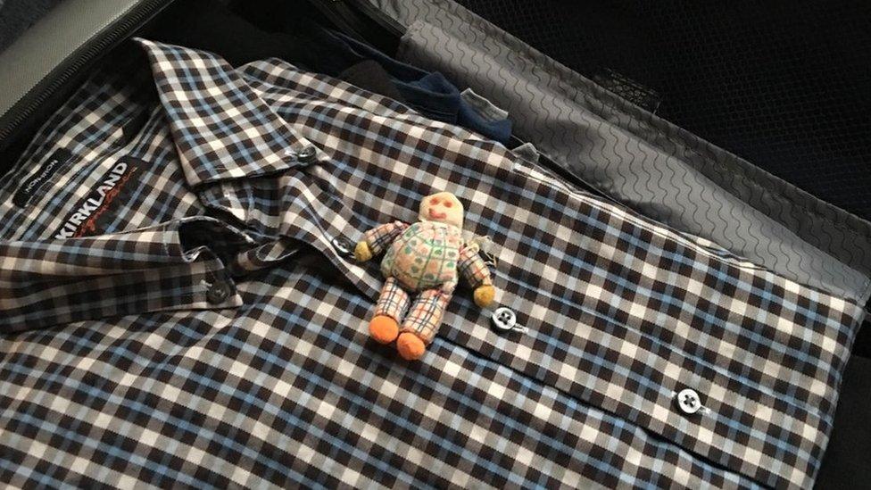 An image of a doll packed in a suitcase on top of a shirt.