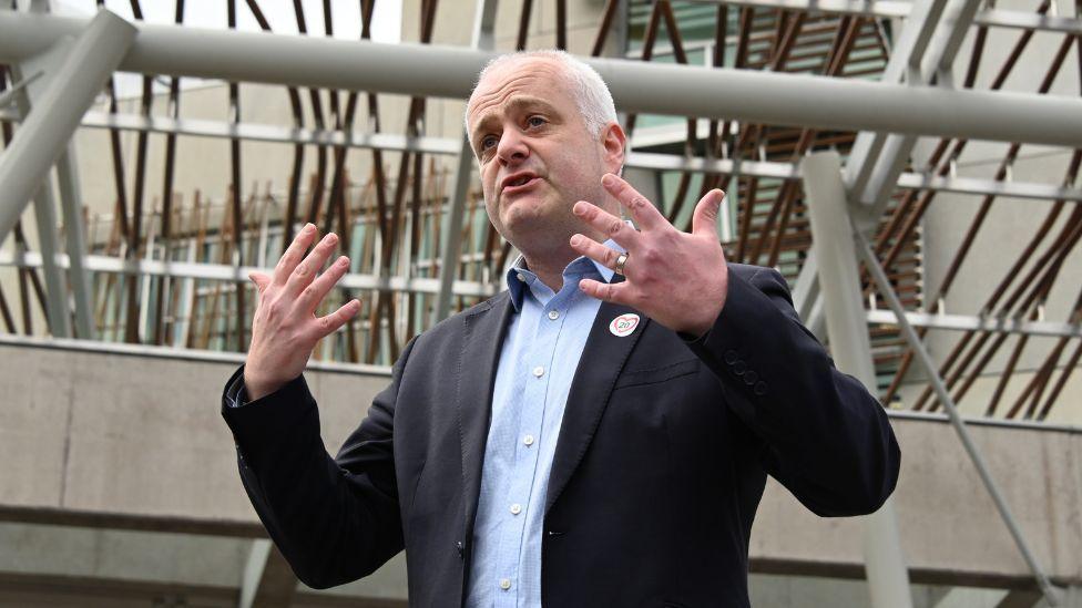 Mark Ruskell in a dark suit and open-neck start standing outside the Scottish Parliament building