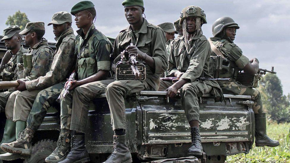 M23 fighters in fatigues and boots and holding guns sit on the back black of a pick-up truck - taken in 2023