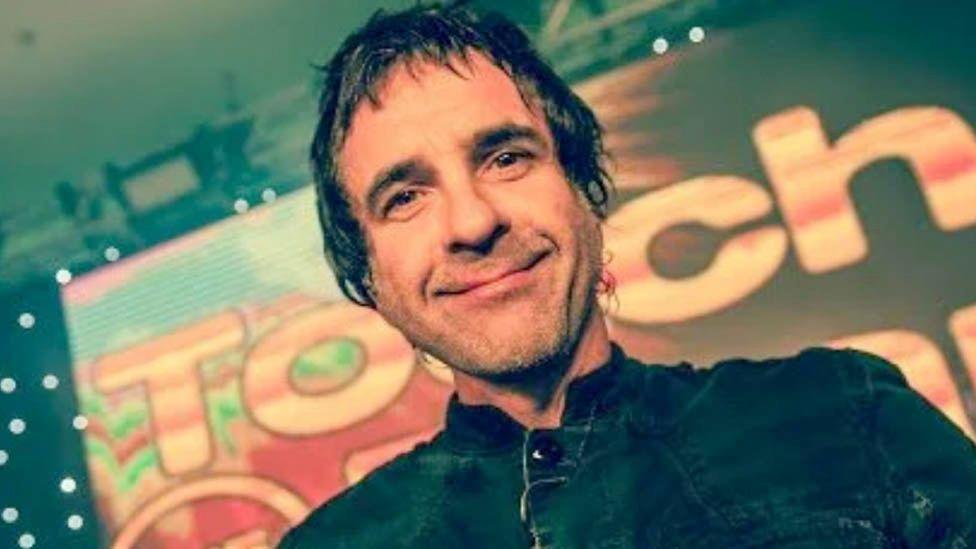 Kev Schools looking down slightly, smiling at the camera. He has short dark hair and stubble and wears a black jacket