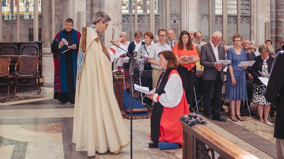 Woman in red and white vestments kneels before a bishop