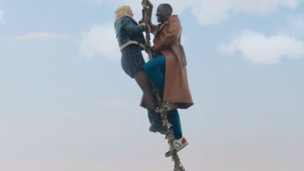 The Doctor, wearing a long brown coat and coloured sneakers, climbs up a rope ladder with his assistant, Ruby