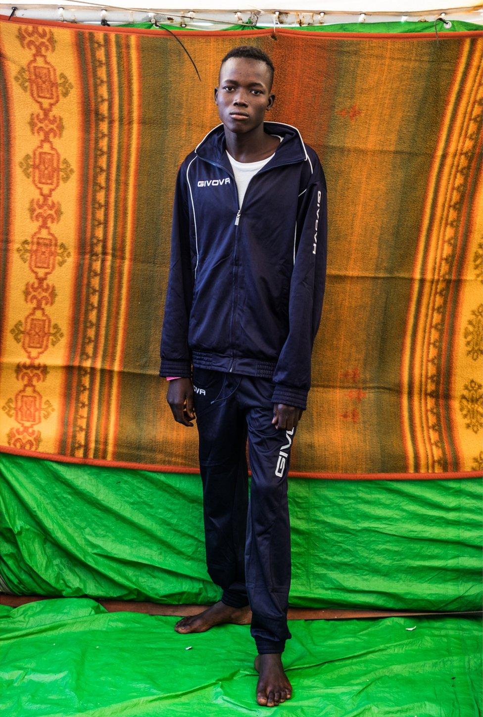 Abdusalam Adam wears a tracksuit and stands in front of a coloured blanket