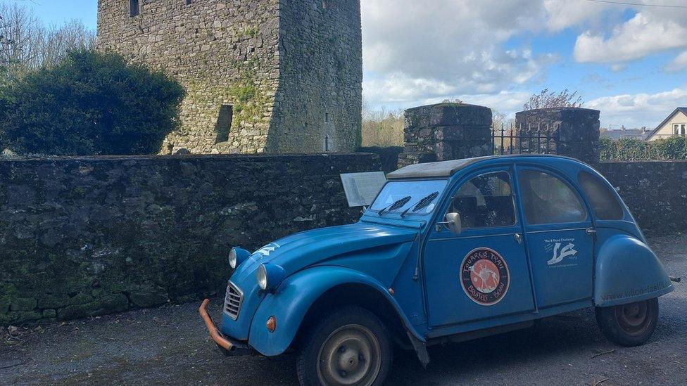 Blue 2CV parked in front of a stone monument or tower