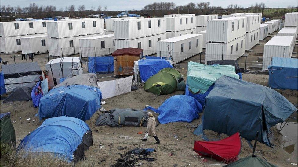 A migrant walks among tents in a makeshift camp as containers are put into place to house several hundred migrants living in what is known as the "Jungle"