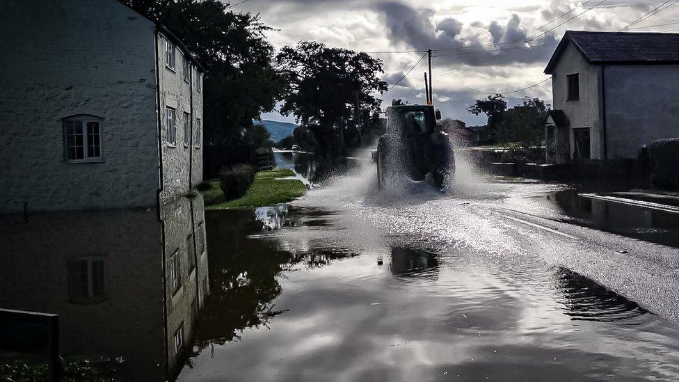 tractor is driving through water, with spray coming from is tyres, on a flooded road through a village