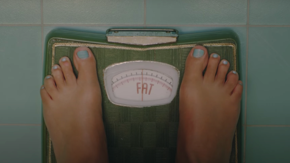 A screenshot from the music video showing bathroom scales that read FAT