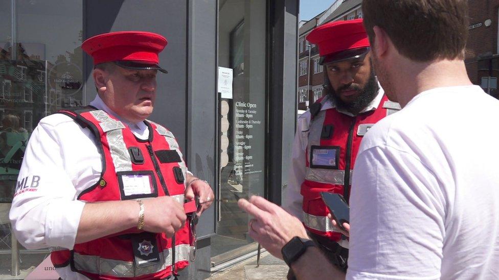 Two Bobbies, wearing red vests, red hats and white shirts, help a member of the public in London.